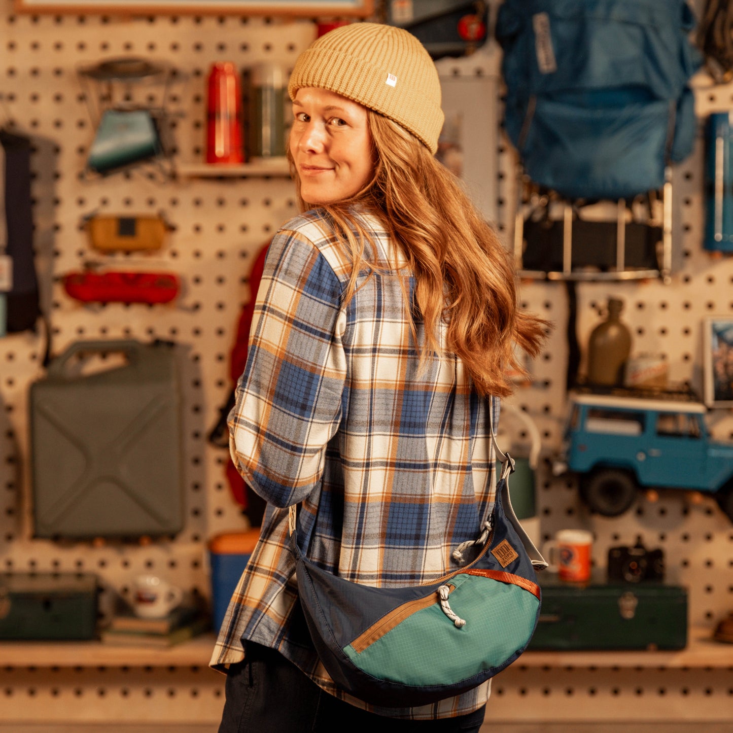 General shot of Person wearing a plaid shirt and beanie with a green bag, standing in a store with shelves and products.