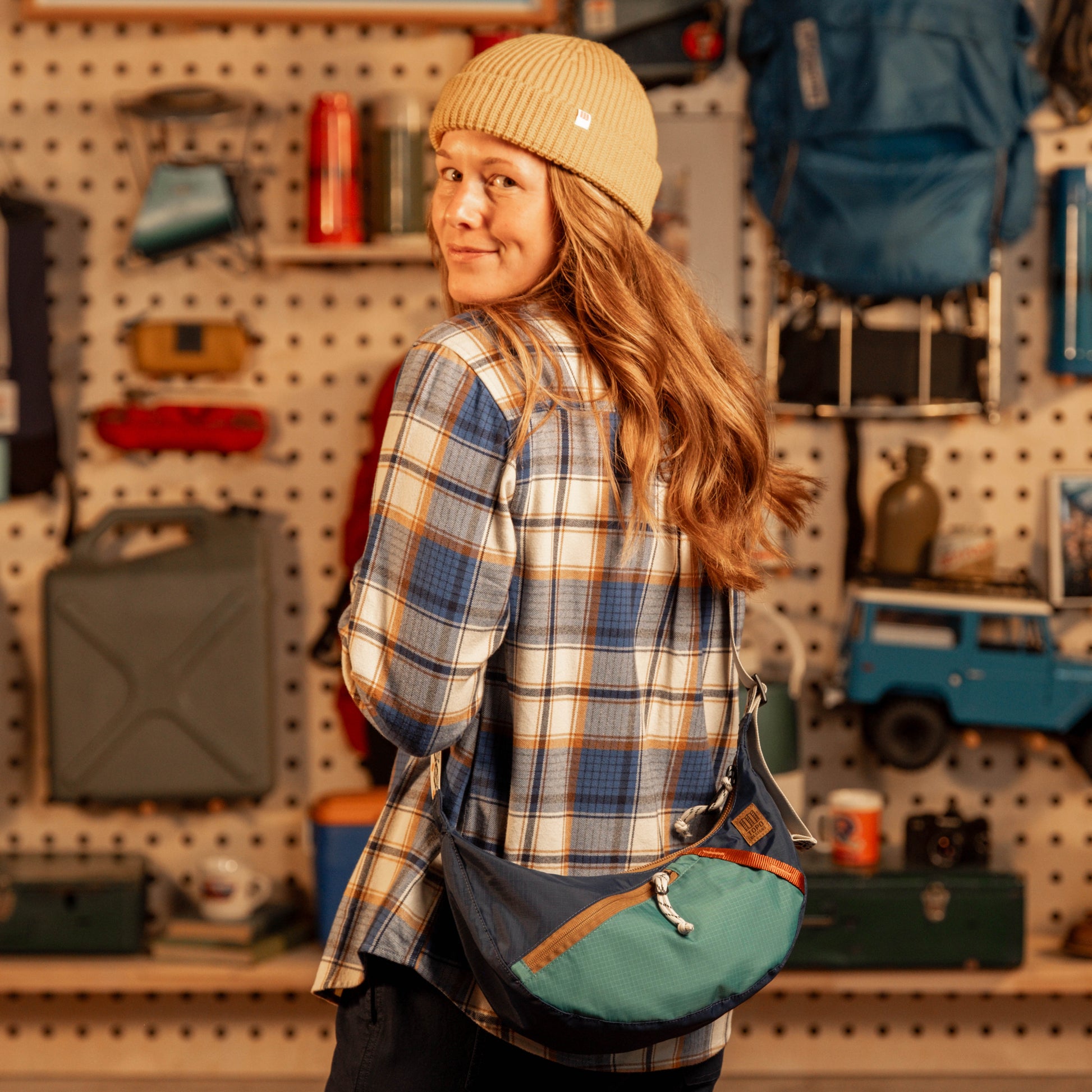 General shot of Person wearing a plaid shirt and beanie with a green bag, standing in a store with shelves and products.